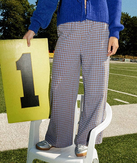 Nika steht auf einem weißen Campingstuhl auf einem Spielfeld und hält eine gelbe Tafel mit einer großen schwarzen Eins darauf in der Hand. Sie trägt eine braun-blau karierte Hose, einen blauen Cardigan und silberfarbene Sneaker; Zu Hosen und Röcke Damen gehen