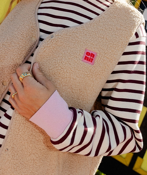 Nika trägt einen gestreiften Sweater in Cremeweiß und Bordeauxrot und darüber eine beigefarbene Fleece-Weste; Zu Jacken Damen gehen