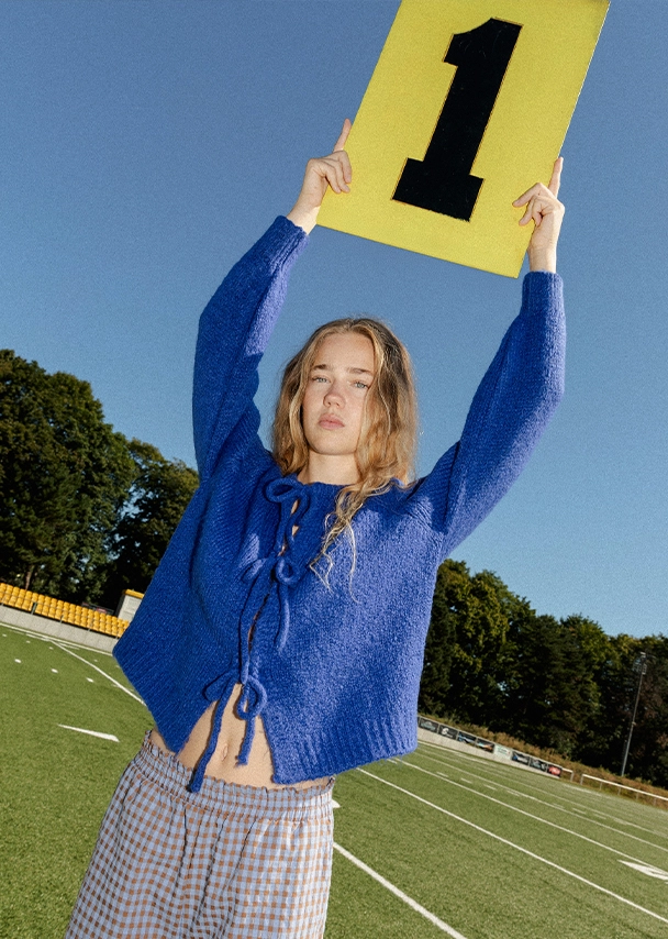 Nika steht auf einem Spielfeld und hält eine Nummerntafel über ihren Kopf. Sie trägt einen blauen Strickcardigan und eine braun-blau karierte Hose; Zur abgebildeten Hose gehen