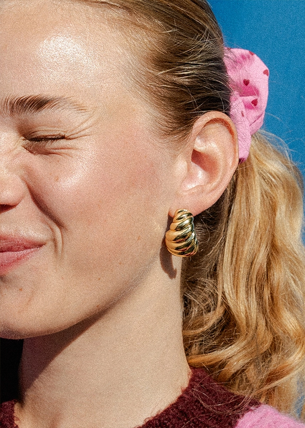 Nika zieht eine Grimasse mit ihrem Gesicht und trägt in ihrem Haar ein rosafarbenes Scrunchie mit kleinen roten Herzen und an den Ohren große goldene Creolen; Zu Ohrringe Damen gehen