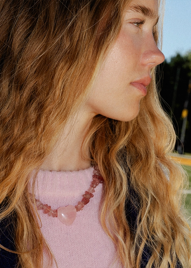 Nika trägt einen rosafarbenen Strickpullover und eine Kette aus kleinen Edelsteinen und einem großen Herzanhänger in Rosa; Zu Schmuck und Accessoires Damen gehen