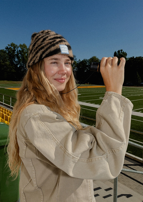 Nika steht auf der Tribüne eines Footballstadions und hält ein Fernglas in der Hand. Sie trägt eine beigefarbene Jacke und eine Mütze mit braun-schwarzem Zebramuster; Zur abgebildeten Mütze gehen