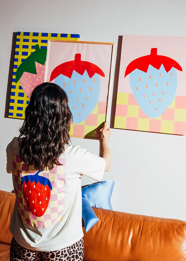 Julia steht vor einer Wand, an der Erdbeer-Bilder hängen. Sie trägt ein cremefarbenes T-shirt mit großem Backprint einer rot-blauen Erdbeere auf einem rosa-gelben Schachbrettmusterhintergrund sowie eine Leopardenprinthose. In den Händen hält sie ein eingerahmtes, farbenfrohes Poster in Rosa, Blau, Gelb, Orange und Pfirsichfarben mit großem Erdbeerdruck; Zum abgebildeten T-Shirt gehen
