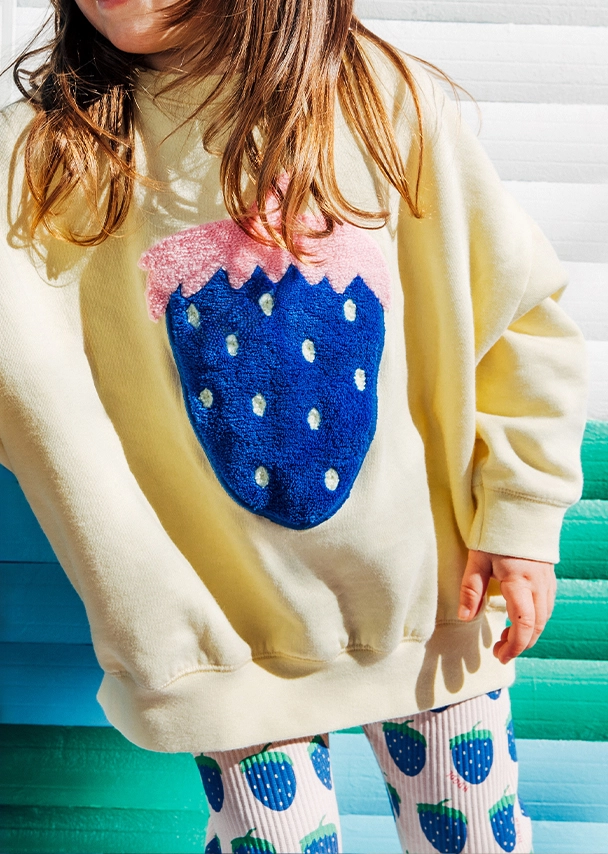 Ein kleines Mädchen steht vor einer bunten Wand. Der Fokus liegt auf ihrem hellgelben Sweater mit großem, kuscheligem Erdbeermotiv in Blau. Dazu trägt sie eine passende Leggins mit blauem Erdbeermuster; Zum abgebildeten Kids-Sweater gehen