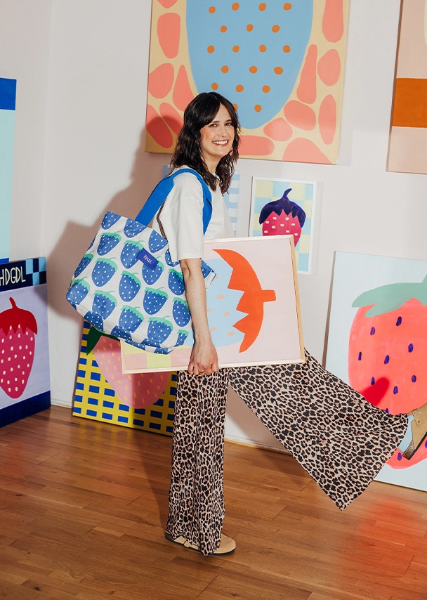 Julia steht lachend vor bunten Leinwänden mit großen Erdbeermotiven. Sie trägt ein hellgelbes T-Shirt und eine lockere Leopardenprinthose mit hellbraunen Birkenstock-Schuhen. Über ihre Schulter trägt sie eine blaue Tragetasche mit Erdbeermuster. Unter ihrem Arm trägt sie ein eingerahmtes Poster mit blau-orangenem Erdbeermotiv; Zur abgebildeten Tragetasche gehen