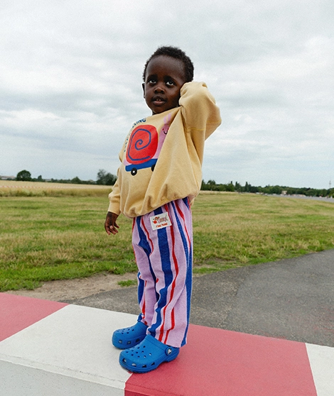Ein Kind steht draußen auf einem Betonblock und trägt einen gelben Sweater mit buntem Schnecken-Print und eine lilafarbene Frotteehose mit blauen und roten Streifen und kleinem Dackel-Patch am Bund; Zum abgebildeten Sweater gehen