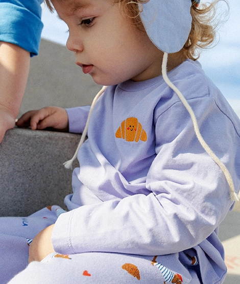 Ein kleines Kind sitzt draußen auf einem Betonblock und trägt einen lilafarbenen Longsleeve und eine lilafarbende Hose mit bunten Croissants