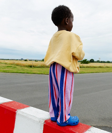 Ein Kind steht draußen auf einem Betonblock mit dem Rücken zu uns gewandt. Es trägt einen gelben Sweater und eine lilafarbene Frotteehose mit blauen und roten Streifen