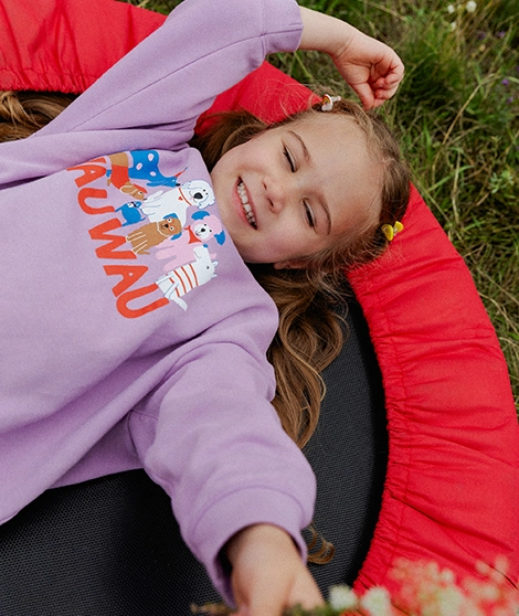 Ein Mädchen liegt auf einem kleinen Trampolin. Es trägt einen lilafarbenen Frottee-Pullover mit großem, buntem Hundeprint mit ?WAU WAU?-Aufschrift in Rot