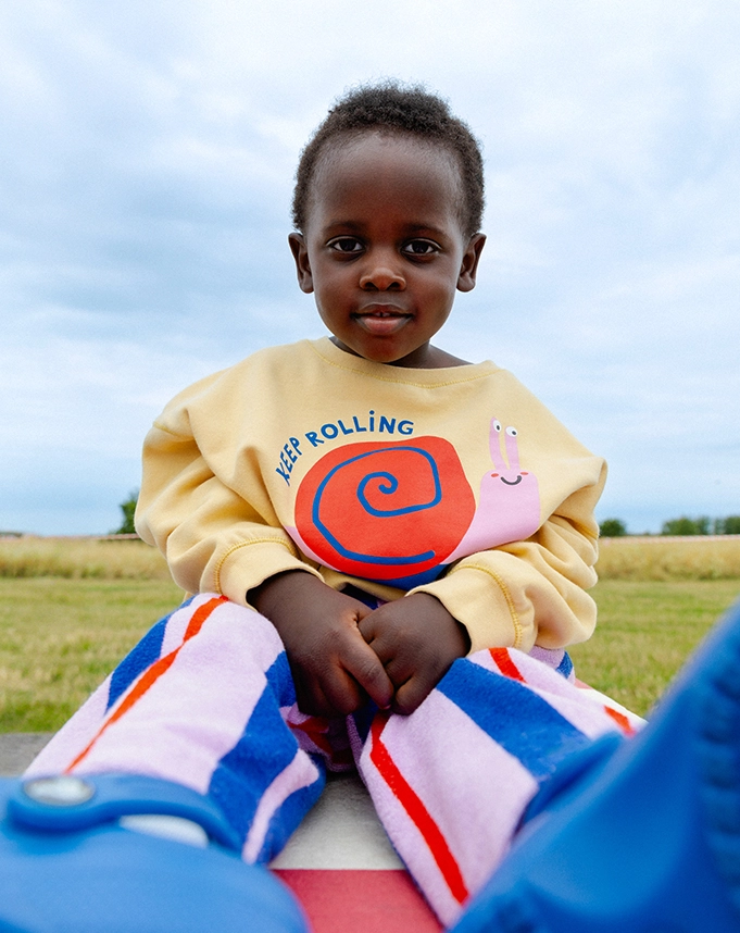 Ein Kind sitzt auf einer Rasenfläche und trägt einen gelben Sweater mit buntem Schnecken-Print und ?KEEP ROLLINGg?-Aufschrift. Dazu trägt es eine lilafarbene Hose aus Frottee mit blauen und roten Streifen; Zum abgebildeten Sweater gehen
