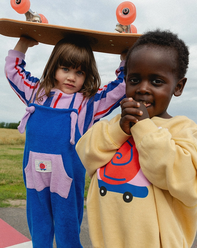 Zwei Kinder stehen nebeneinander auf einer Rasenfläche. Das linke Kind trägt einen blauen Overall über einen lila-rot-blau-gestreiften Pullover aus Frottee und hält ein Skateboard über dem Kopf. Das zweite Kind trägt einen gelben Sweater mit buntem Schnecken-Print und ?KEEP ROLLINGg?-Aufschrift; Zum abgebildeten Overall gehen