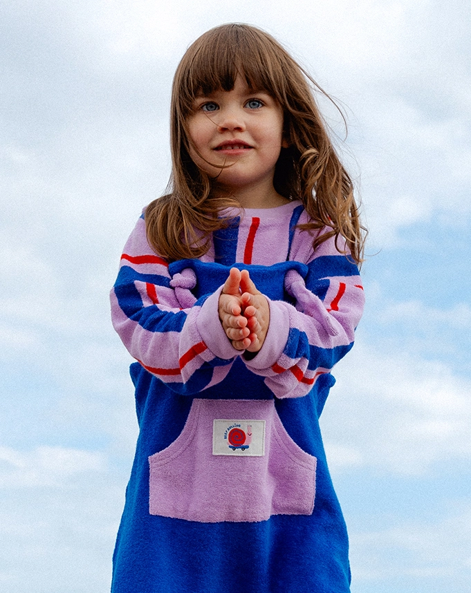 Ein kleines Mädchen steht unter blauem Himmel und trägt einen blau-lilafarbenen Overall aus Frottee und darunter einen lilafarbenen Frottee-Pullover mit blauen und roten Streifen. Auf der Bauchtasche des Overalls ist ein kleines, farbenfrohes Patch mit einer Skateboard fahrenden Schnecke gestickt; Zum abgebildeten Overall gehen