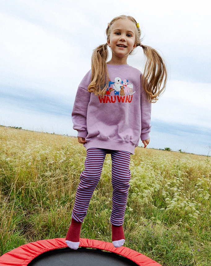 Ein Mädchen hüpft auf einem kleinen Trampolin vor einer grünen Wiese. Es trägt einen lilafarbenen Frottee-Pullover mit großem, buntem Hundeprint mit ?WAU WAU?-Aufschrift in Rot und eine dazu passende Frottee-Hose mit roten und blauen Streifen; Zum abgebildeten Pullover gehen