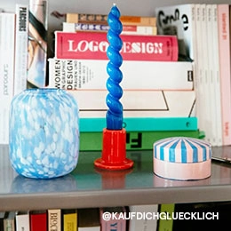 Ein Regalbrett, auf dem eine blau marmorierte Vase, eine blaue gedrehte Kerze in einem roten Kerzenhalter und einer kleinen gestreiften Schmuckbox stehen. Das Bild ist vom Instagram Account von @KAUFDICHGLUECKLICH; Zur abgebildeten Vase gehen