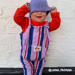 Auf dem Bild sieht man ein Baby, welches an einer Wand steht und sich eine liane Mütze über die Augen zieht. Dazu trägt es ein rotes Longsleeve und einen gemusterten Overall aus unserer Kids-Kollektion. Das Bild ist vom Instagram Account von @JANA.PAPANA; Zum abgebildeten Overall gehen.