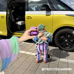 Zwei kleine Kinder umarmen sich vor einem gelben Bus. Sie tragen bunte Mützen und das vordere Kind trägt einen gestreiften Overall in Lila, Blau und Rot und darüber eine grüne Jacke. Eine Hand hält ein lilafarbenes, schimmerndes Osterei so ins Bild, dass das Gesicht des hinteren Kindes verdeckt wird; Zum abgebildeten Kids-Overall gehen