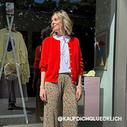 Silvana steht vor dem KAUF DICH GLÜCKLICH-Schaufenster in Berlin und trägt ein weißes T-Shirt, einen knallroten Cardigan und eine bunt gemusterte Hose; Zum abgebildeten Cardigan gehen