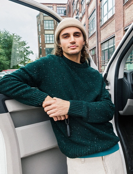 Andris lehnt lässig an einer offenen Autotür. Er trägt einen melierten Strickpullover in Dunkelgrün und eine beigefarbene Rippstrickmütze; Zur Herren New In Seite gehen