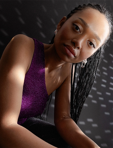 Julia ist in Nahaufnahme zu sehen und blickt direkt in die Kamera. Der Hintergrund ist dunkel und sie trägt ein glitzerndes one-shoulder Top in lila, Zu unseren Festive Essentials gehen